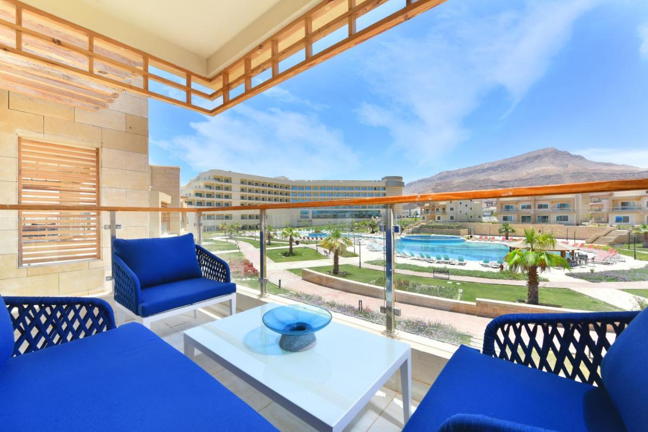 Tolip Resort El Galala Majestic Айн-Сохна Экстерьер фото The photo showcases a balcony view featuring two blue chairs and a table with a decorative bowl. The balcony overlooks a landscape that includes a swimming pool and manicured lawns. In the background, there are multiple buildings and palm trees, all 