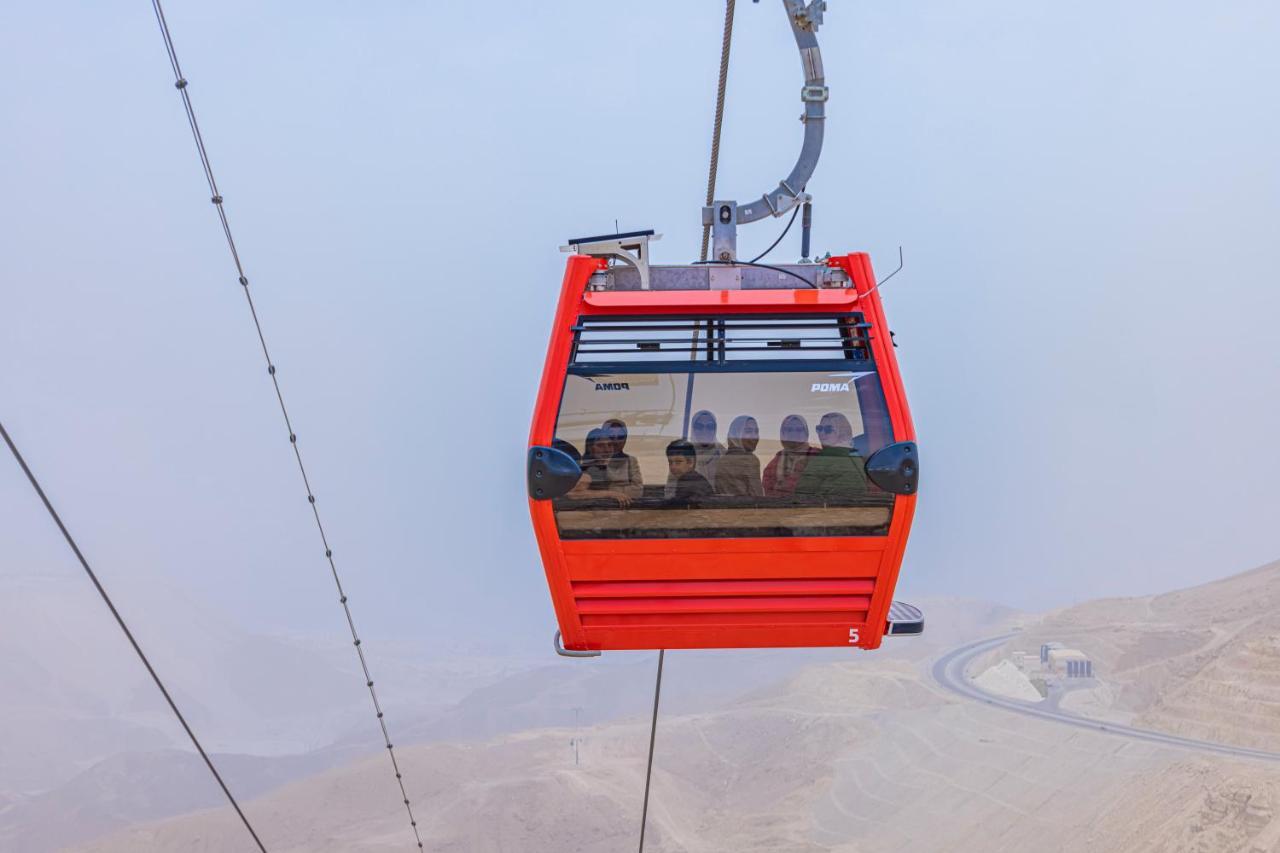 Tolip Resort El Galala Majestic Айн-Сохна Экстерьер фото The photo shows a red gondola lift suspended in the air, traveling along cables. Inside the gondola, a group of people can be seen. The background features a hazy landscape, possibly mountains or hills, indicating an elevated area. The scene conveys 