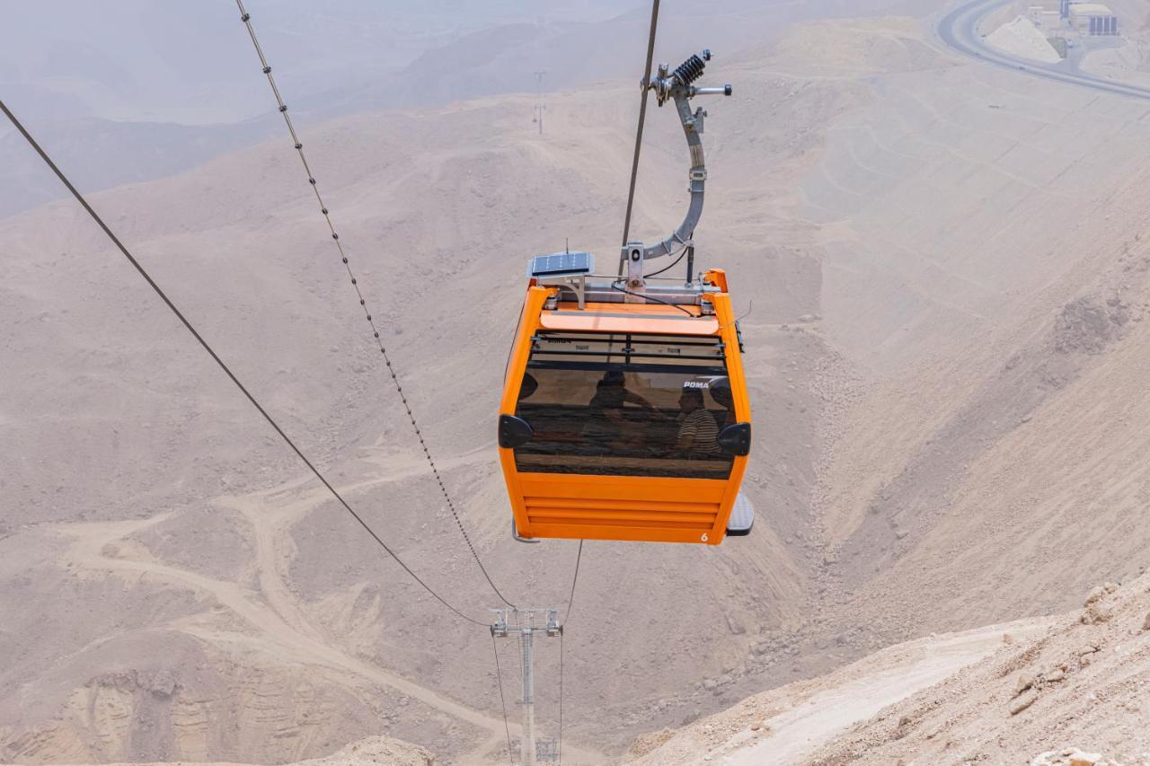 Tolip Resort El Galala Majestic Айн-Сохна Экстерьер фото The photo shows an orange cable car suspended in the air, traveling along a set of cables. It appears to be located in a mountainous or hilly area, with rugged terrain and dry, rocky landscapes visible below. The background features slopes that seem 