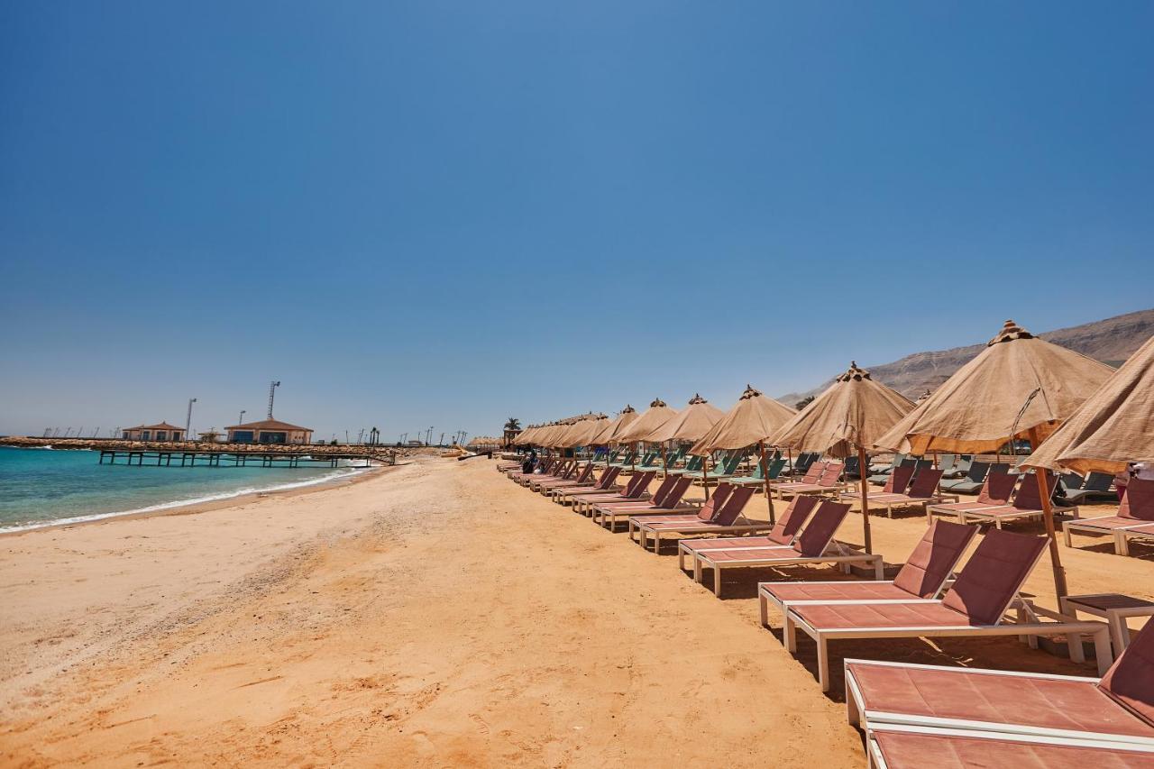 Tolip Resort El Galala Majestic Айн-Сохна Экстерьер фото The photo shows a scenic beach with a stretch of golden sand under a clear blue sky. There are several beach loungers arranged in rows, each accompanied by thatched umbrellas providing shade. In the background, there is a wooden pier extending into t