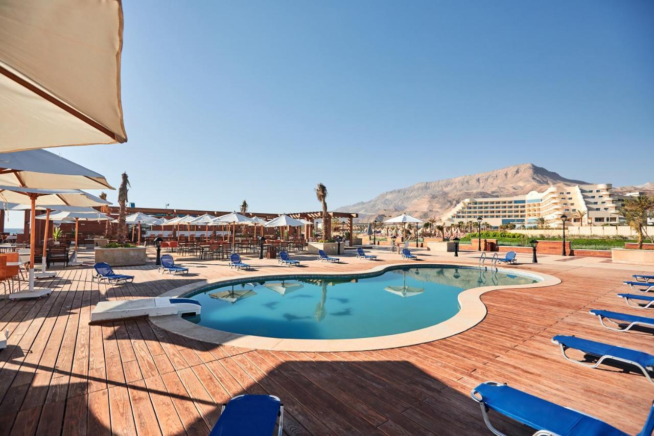 Tolip Resort El Galala Majestic Айн-Сохна Экстерьер фото The photo shows a swimming pool area surrounded by sun loungers and umbrellas. The pool has a unique, curvy shape and is situated on a wooden deck. In the background, there are several buildings, likely part of a resort or hotel, and a mountainous la