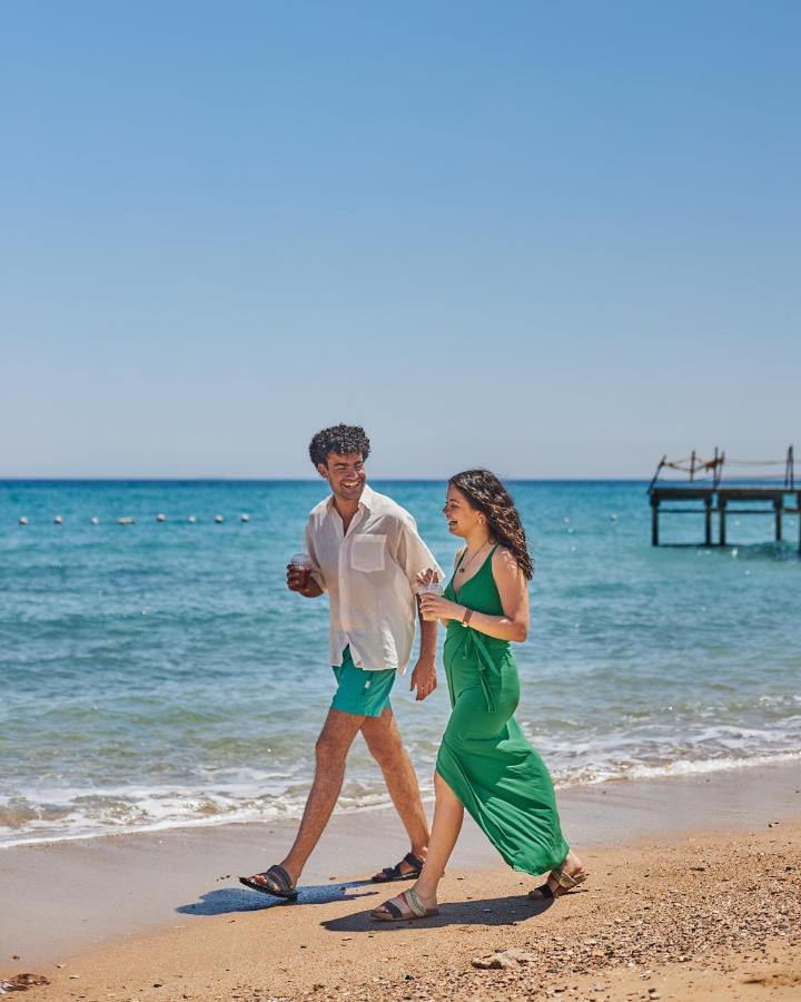 Tolip Resort El Galala Majestic Айн-Сохна Экстерьер фото The photo shows a couple walking along a beach. The man is wearing a white short-sleeved shirt and green shorts, while the woman is dressed in a flowing green dress. They both appear to be enjoying the pleasant weather, with the ocean in the backgrou