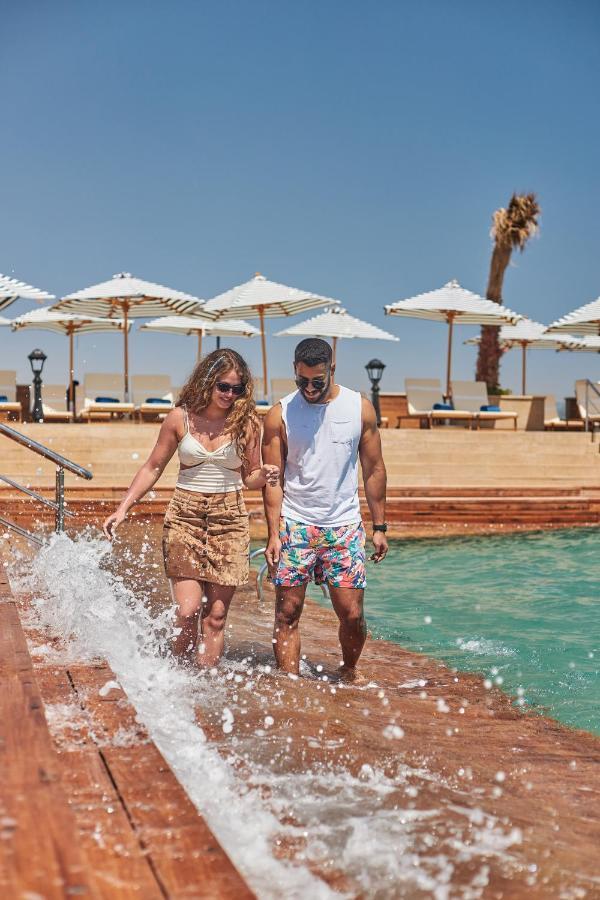 Tolip Resort El Galala Majestic Айн-Сохна Экстерьер фото The photo depicts a couple walking along a poolside area. They are enjoying a sunny day near a swimming pool, with splashes of water around them. The woman is wearing a light-colored top and a patterned skirt, while the man is dressed in a sleeveless