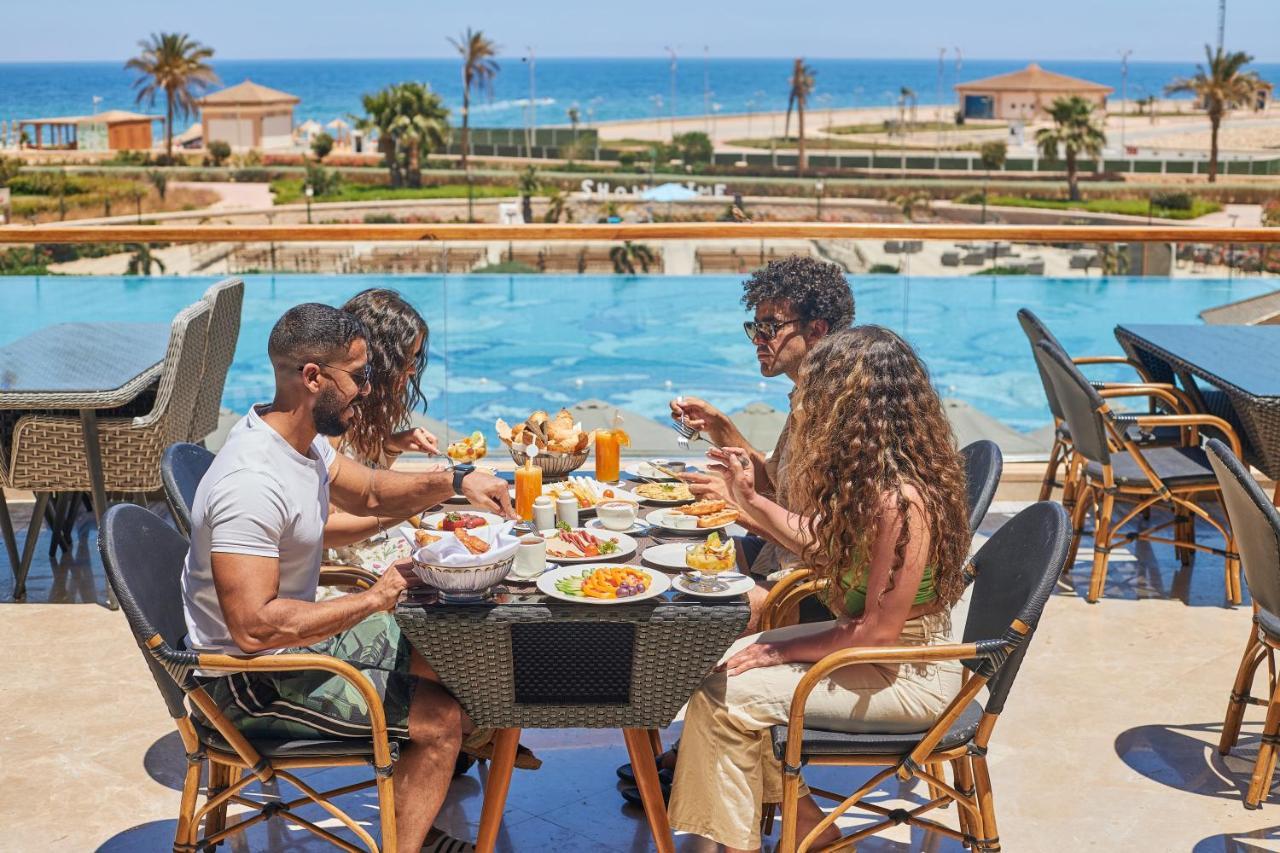 Tolip Resort El Galala Majestic Айн-Сохна Экстерьер фото The photo shows a group of five people enjoying a meal together outdoors, likely at a restaurant or cafe. They are seated at a table with a beautiful view of a swimming pool and the sea in the background. The table is filled with various dishes, and 