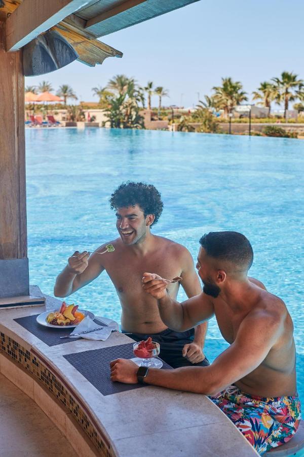 Tolip Resort El Galala Majestic Айн-Сохна Экстерьер фото The photo features two young men enjoying a meal together. They are sitting at a poolside table with a clear blue swimming pool in the background. The men are shirtless, suggesting a casual, relaxed atmosphere, likely in a warm environment. One man i