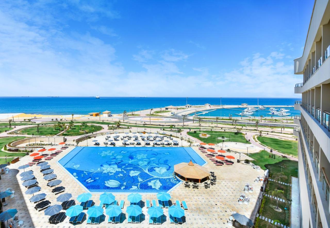 Tolip Resort El Galala Majestic Айн-Сохна Экстерьер фото The photo shows a beautiful outdoor view of a resort area featuring a large swimming pool with a decorative underwater design. Surrounding the pool are numerous sun loungers and umbrellas in a variety of colors. In the background, there is a scenic c