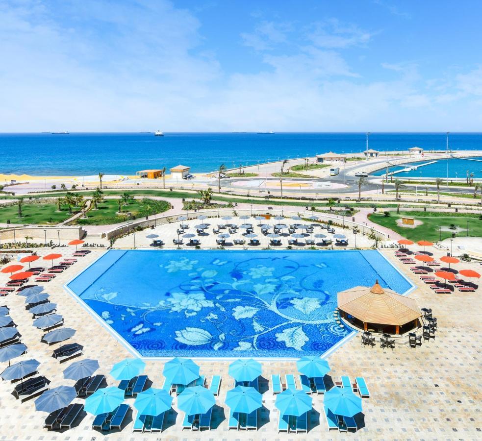 Tolip Resort El Galala Majestic Айн-Сохна Экстерьер фото The photo features a large swimming pool with a decorative design on its surface, surrounded by sun loungers and umbrellas. In the background, there is a view of the sea, with a clear blue sky and a few scattered clouds. The area is well-maintained, 