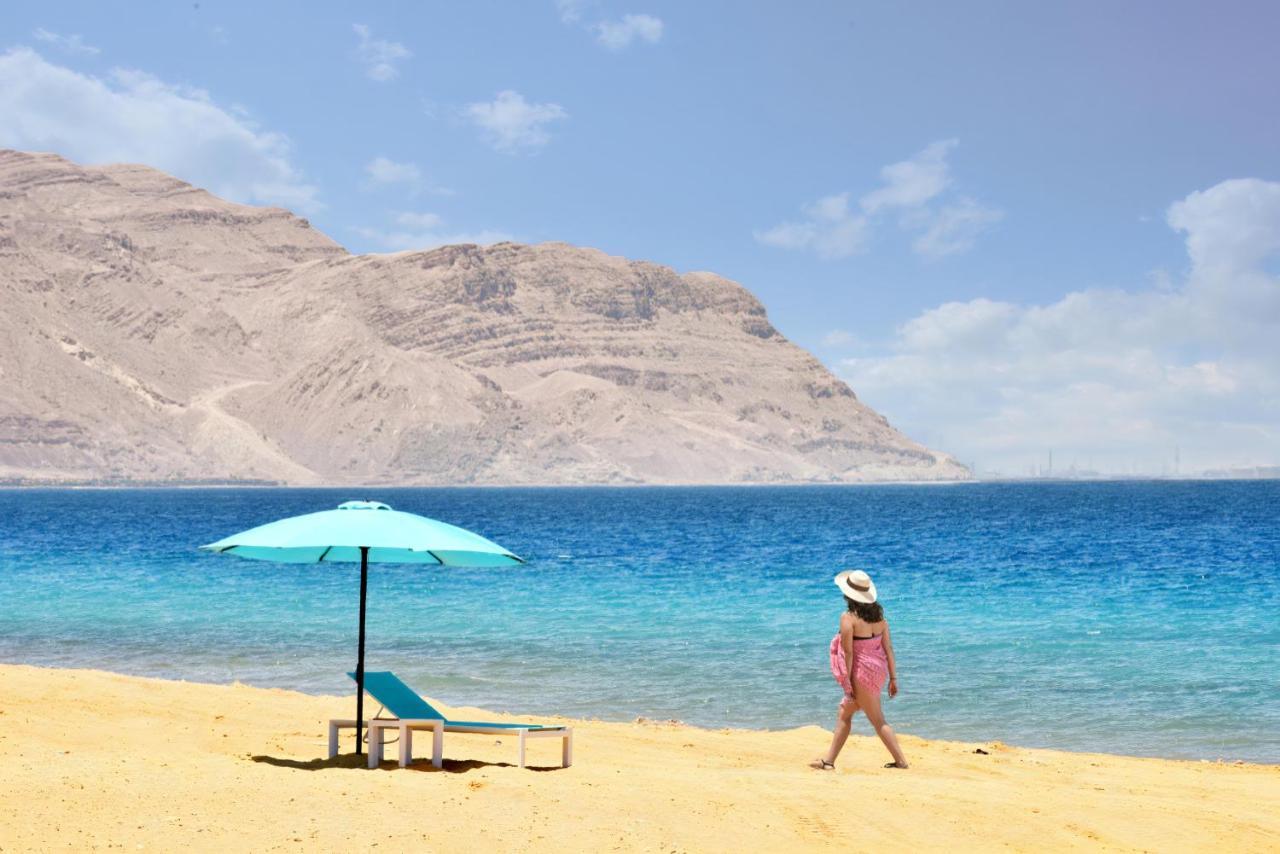 Tolip Resort El Galala Majestic Айн-Сохна Экстерьер фото The photo depicts a serene beach scene. In the foreground, there is a sandy shore with a single beach chair and an umbrella in a light blue color. A person dressed in a pink outfit is walking along the shoreline, enjoying the beach atmosphere. In the